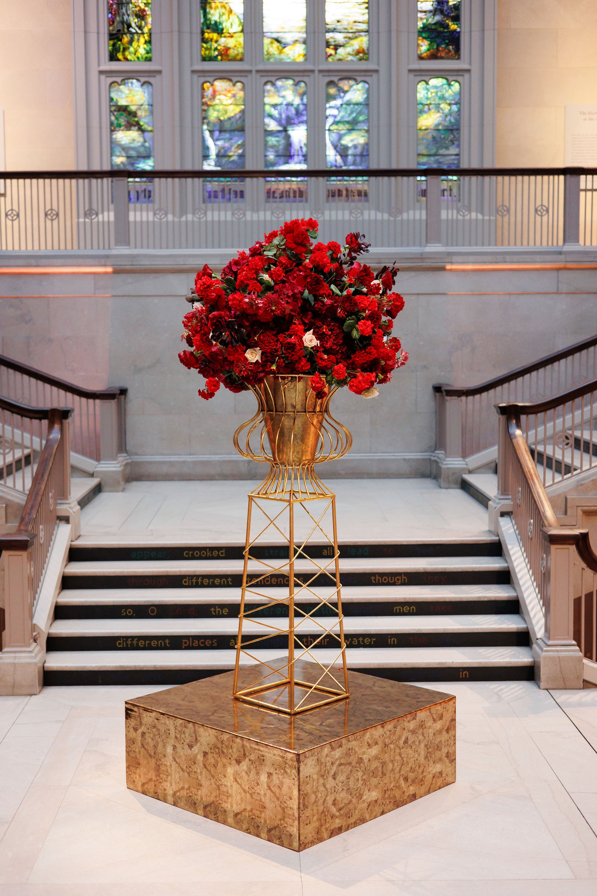 Towering crimson floral urn on a gold pedestal at the foot of the Art Institute of Chicago grand staircase beneath the Tiffany stained-glass windows