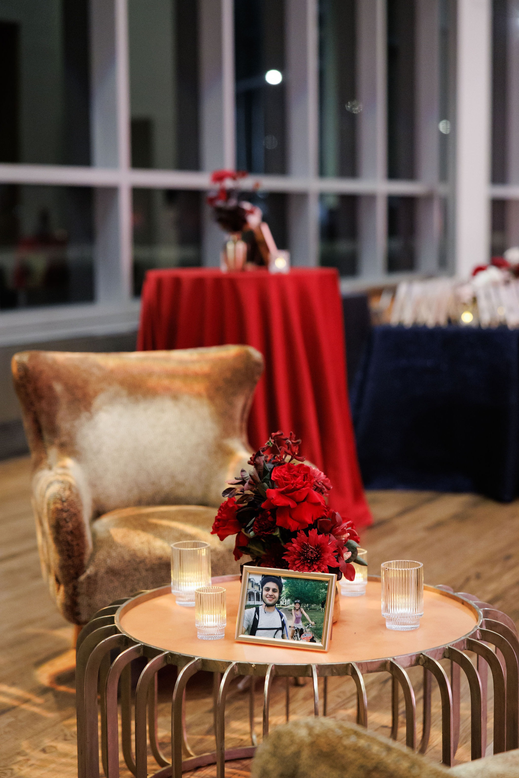 Cocktail-hour lounge with a gold velvet armchair, red roses, votives, and a framed photo of the couple beside crimson and navy cocktail tables