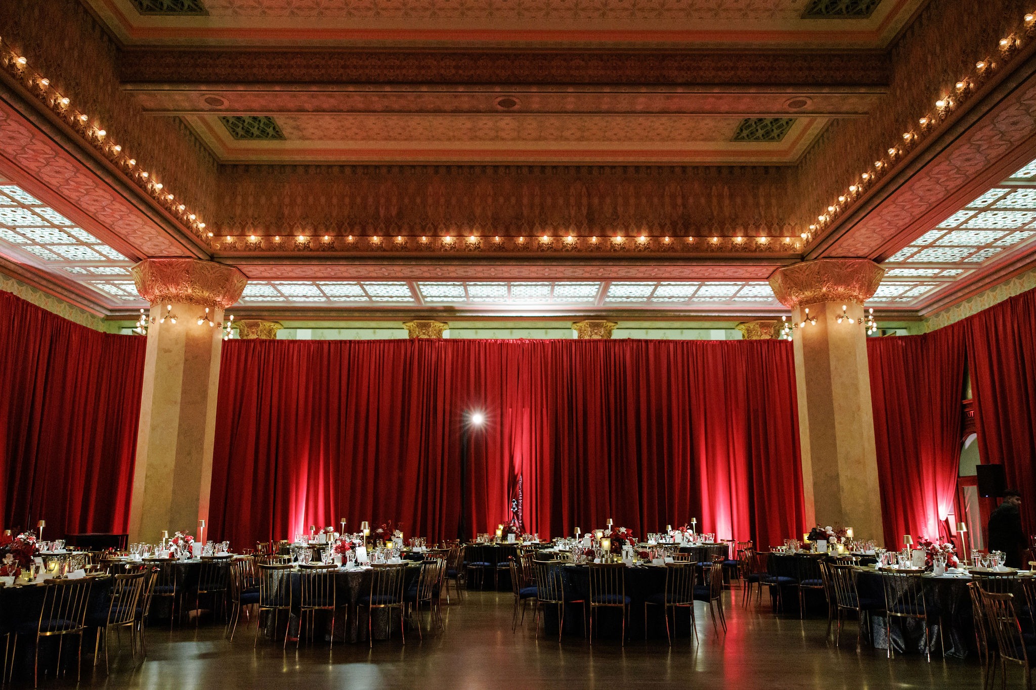 Stock Exchange Trading Room at the Art Institute of Chicago dressed for Lavi and Sid's wedding reception with crimson drapery, gold chairs, and navy-linen dinner tables beneath the stencilled ceiling