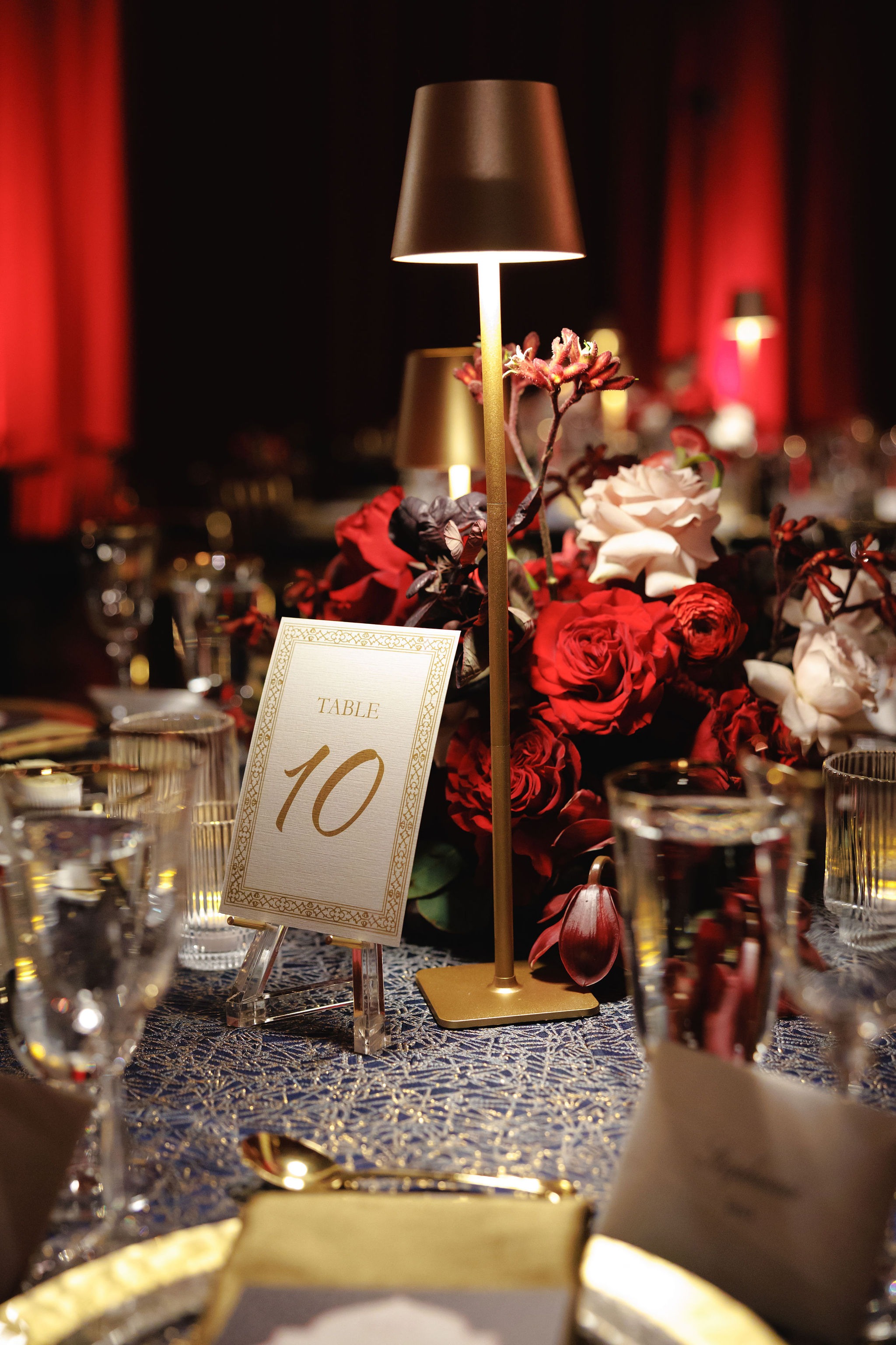 Gold-framed Table 10 card beside a gold cordless lamp and red and blush roses on a royal blue patterned linen