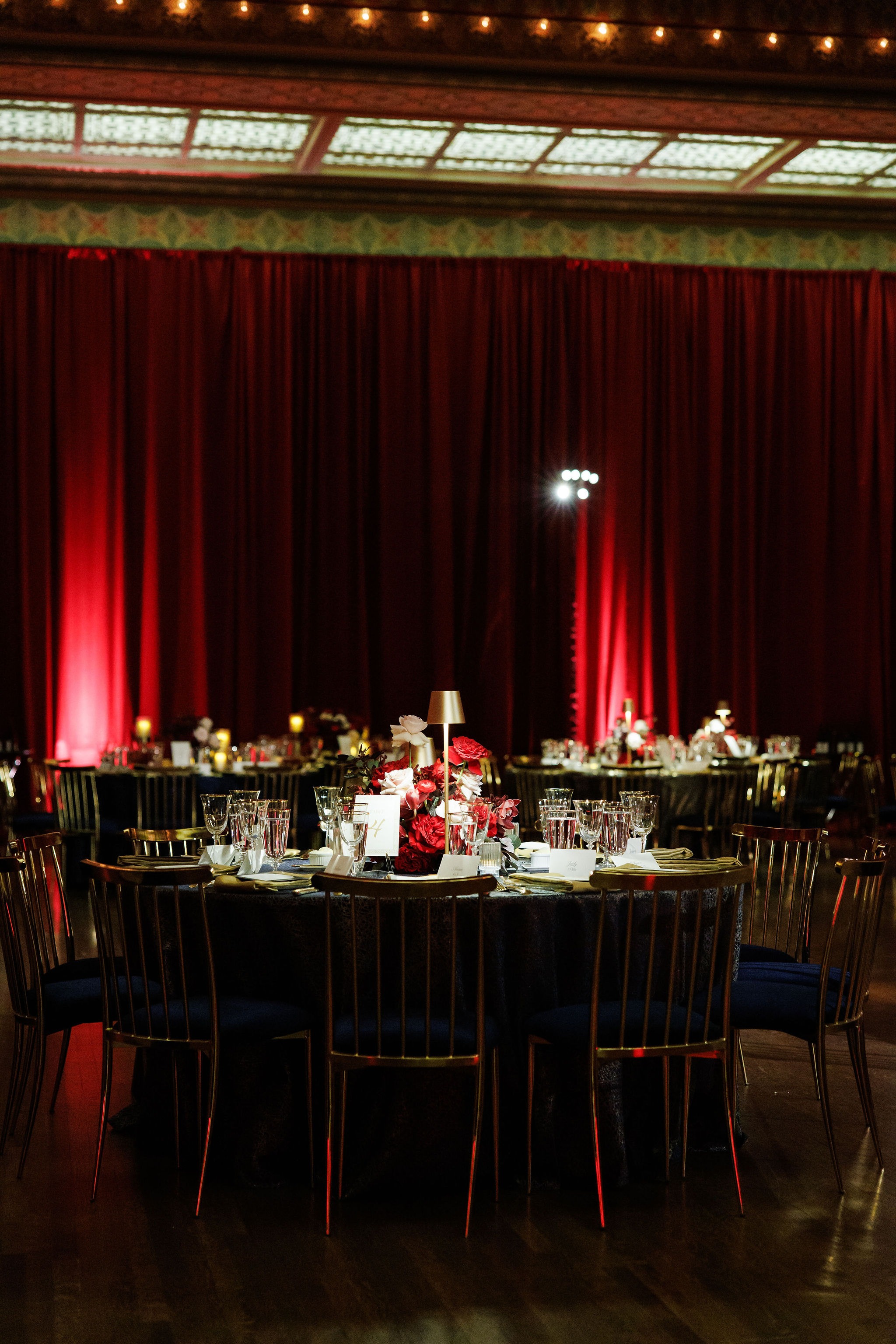 Round dinner table with gold chairs, navy linen, and a crimson centerpiece in front of red-lit drapery in the Stock Exchange Trading Room