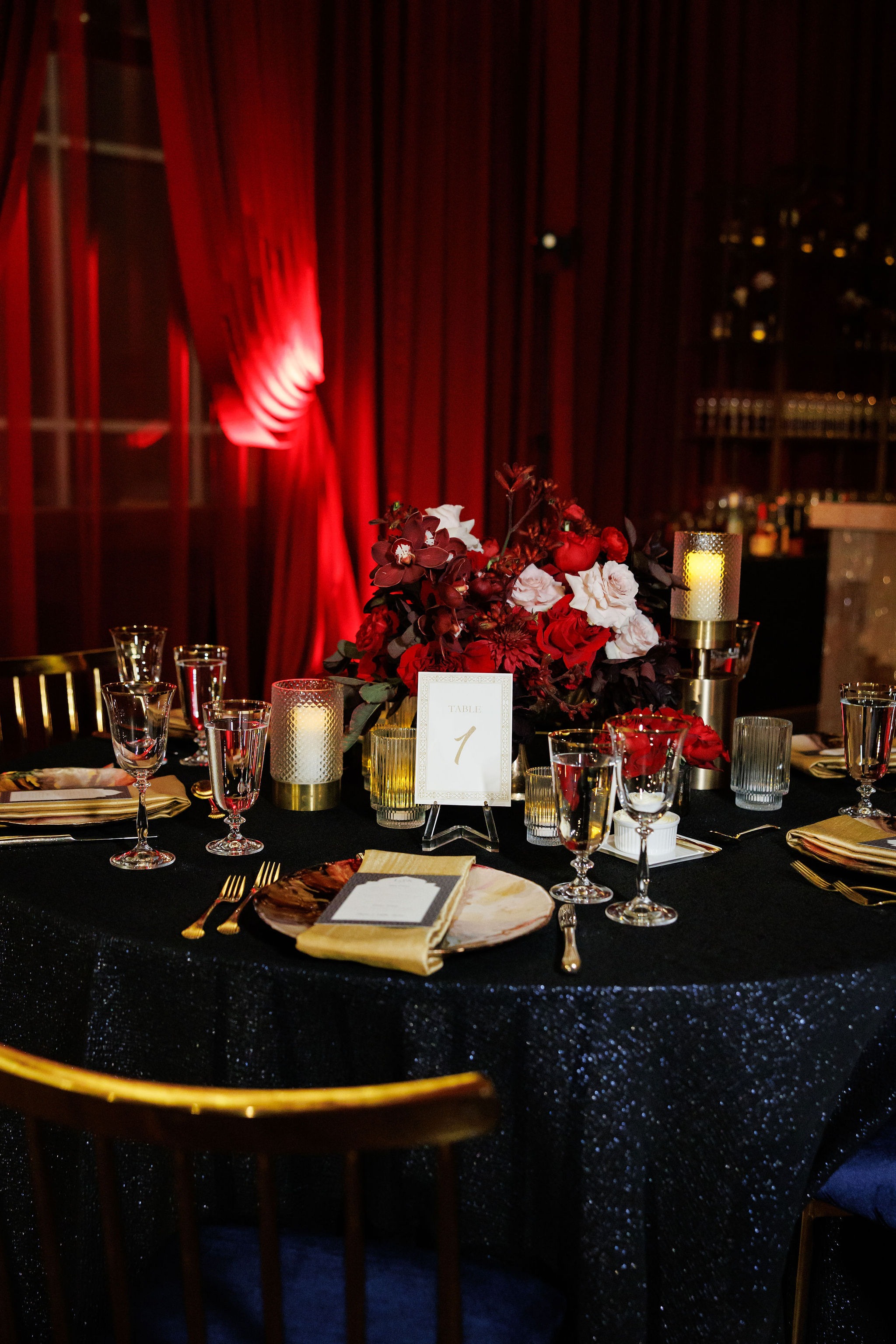 Table 1 place settings with gold flatware, gold napkins, hurricane candles, and a red and blush floral centerpiece on a sequined navy linen