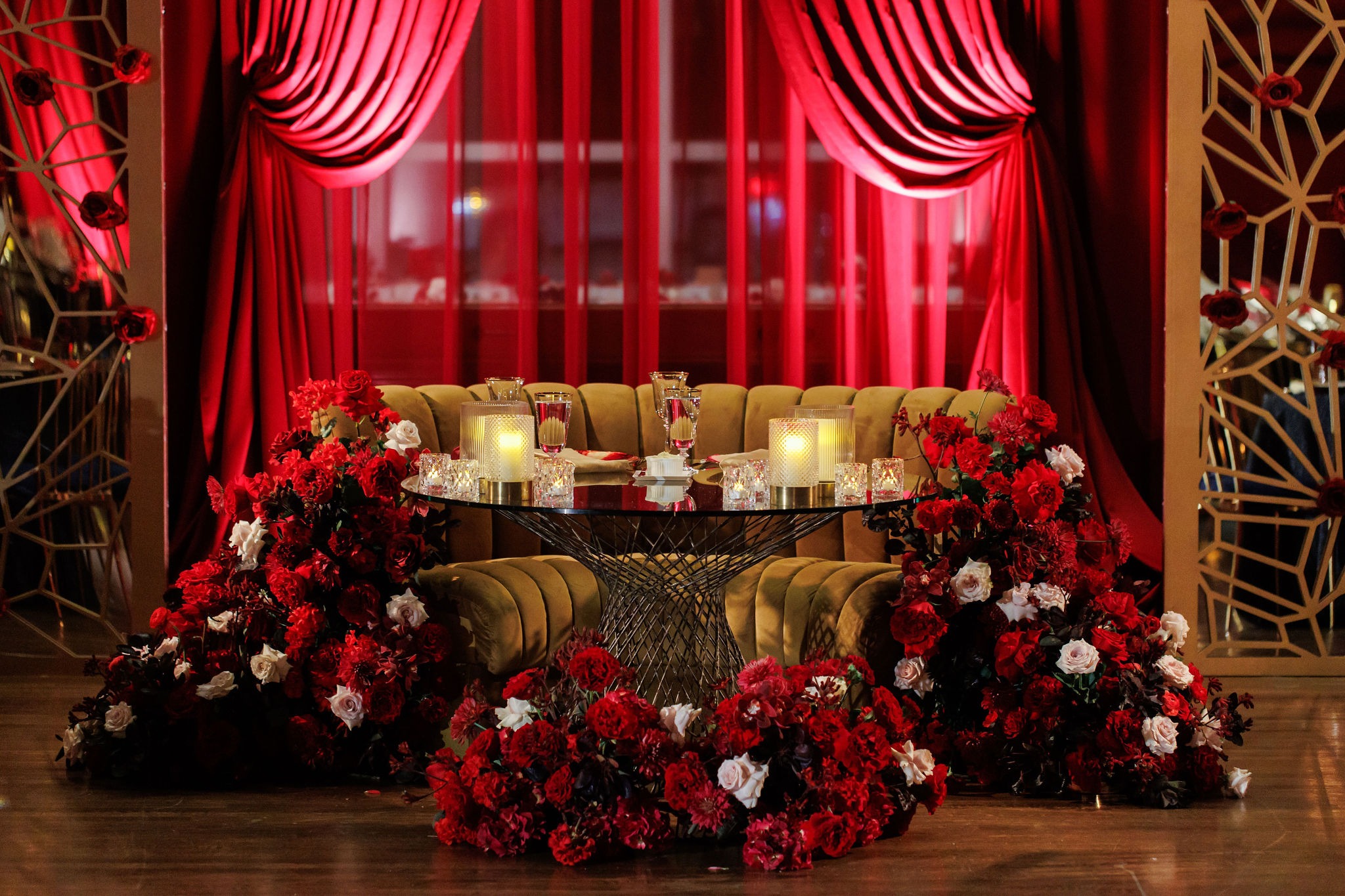 Sweetheart table with a gold velvet settee framed by crimson florals, candlelight, and red draped curtains at Lavi and Sid's reception