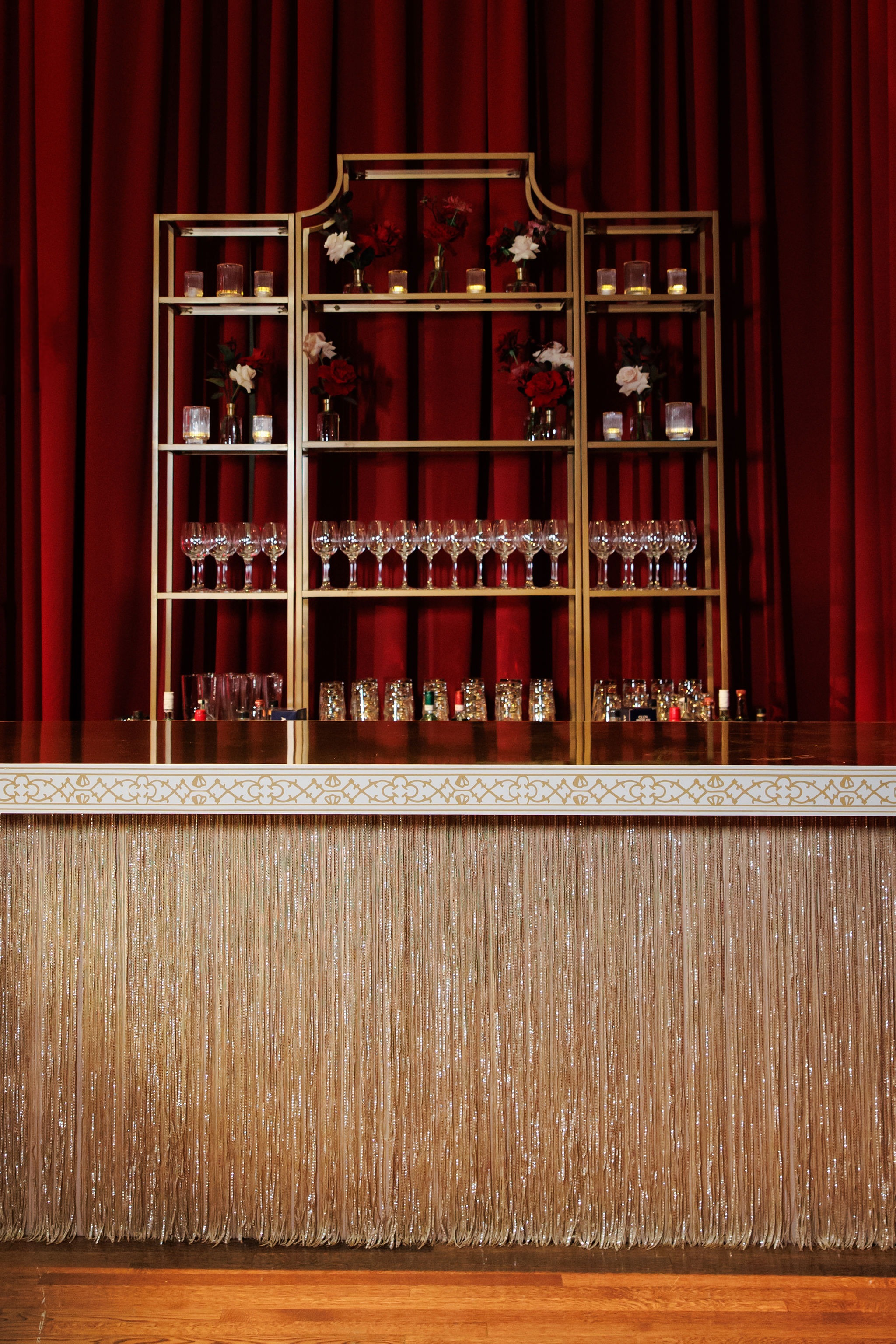 Gold fringed bar with brass back-bar shelving of glassware, votives, and bud vases of red and white roses against crimson drapery