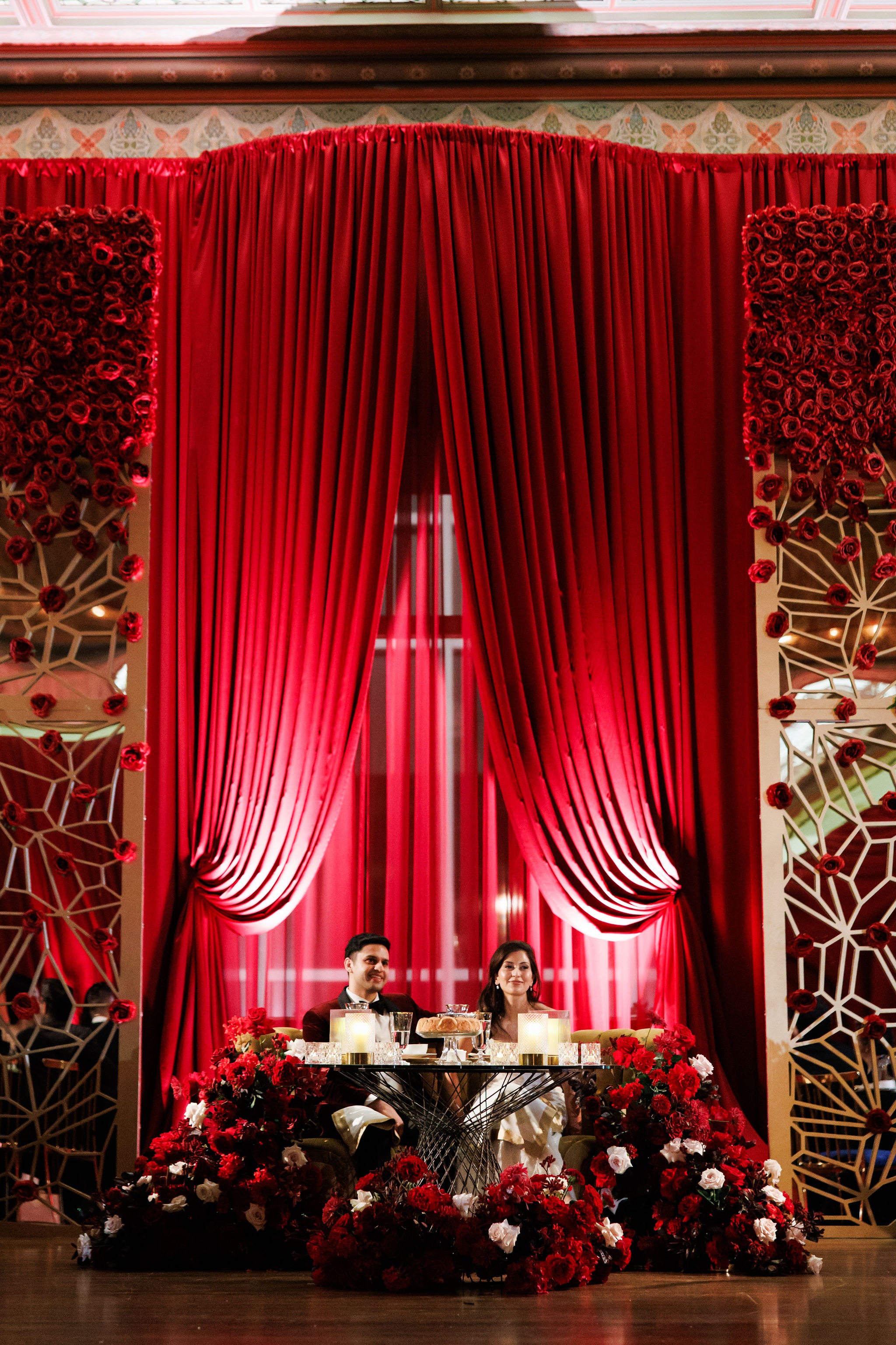 Lavi and Sid seated at their sweetheart table beneath swagged crimson drapery, flanked by gold lattice screens and rose walls