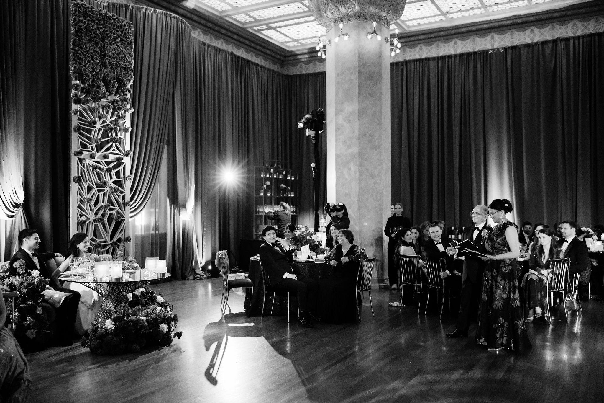 Black-and-white view of toasts in the Stock Exchange Trading Room, with the couple at their sweetheart table and guests looking on