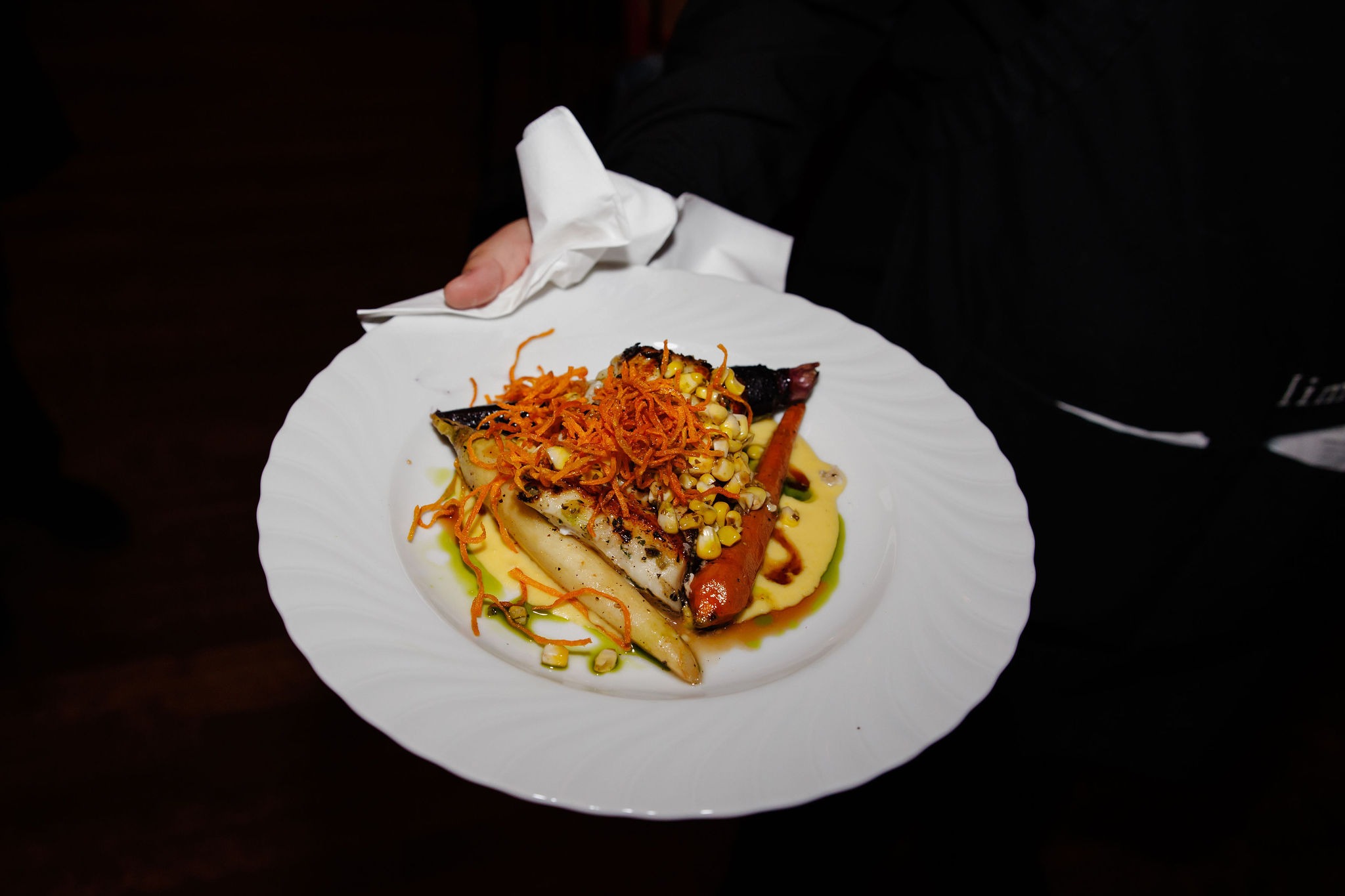 Server presenting a plated entrée of seared fish over purée with roasted carrot, charred corn, and crisp shoestring garnish