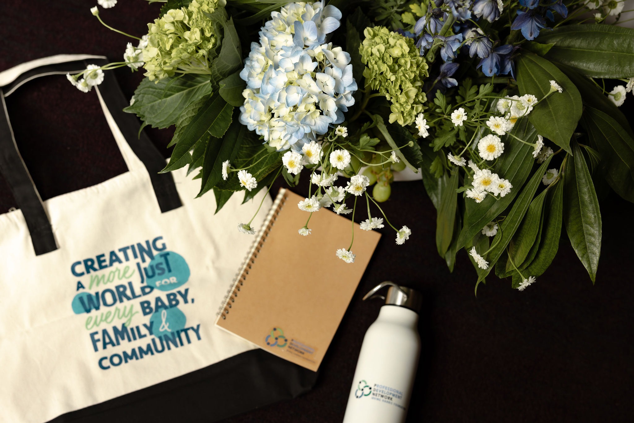 Conference welcome tote, notebook and water bottle beside blue hydrangea and delphinium florals