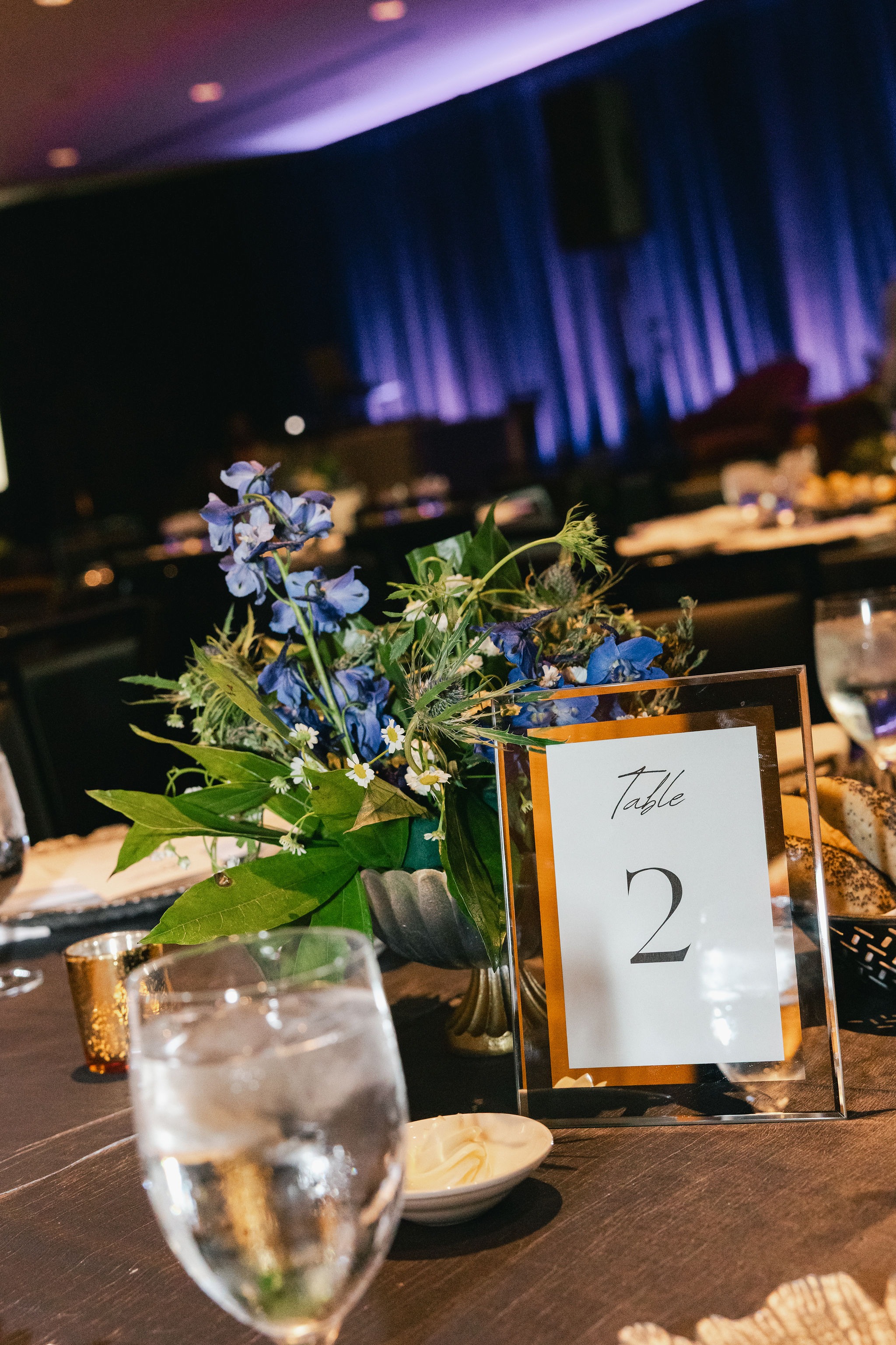 Table number card and blue delphinium centerpiece in a violet-lit conference dining room