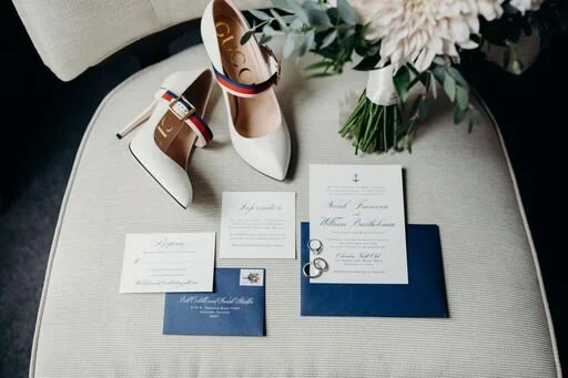 Wedding day flat lay of navy and ivory stationery, rings, bridal heels, and a bouquet on a chair
