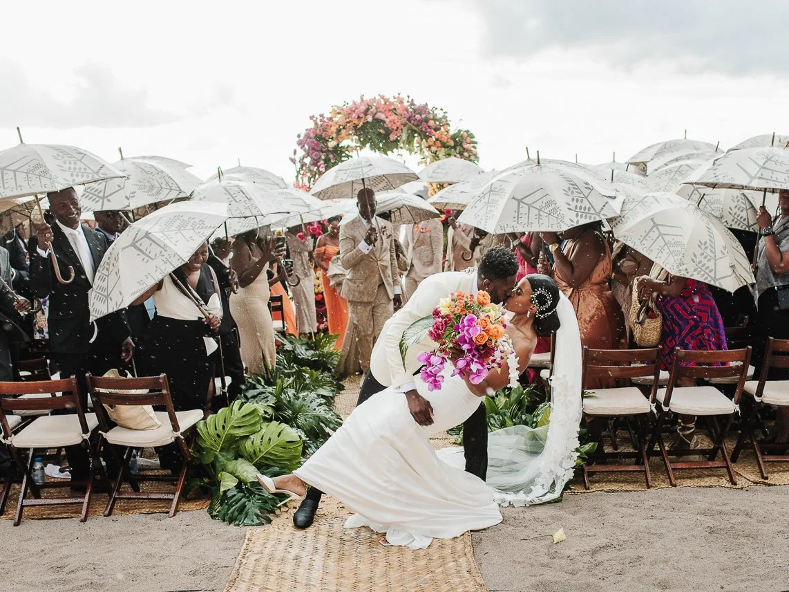 Jean and Jennifer kiss in the rain under guest umbrellas at their seaside ceremony
