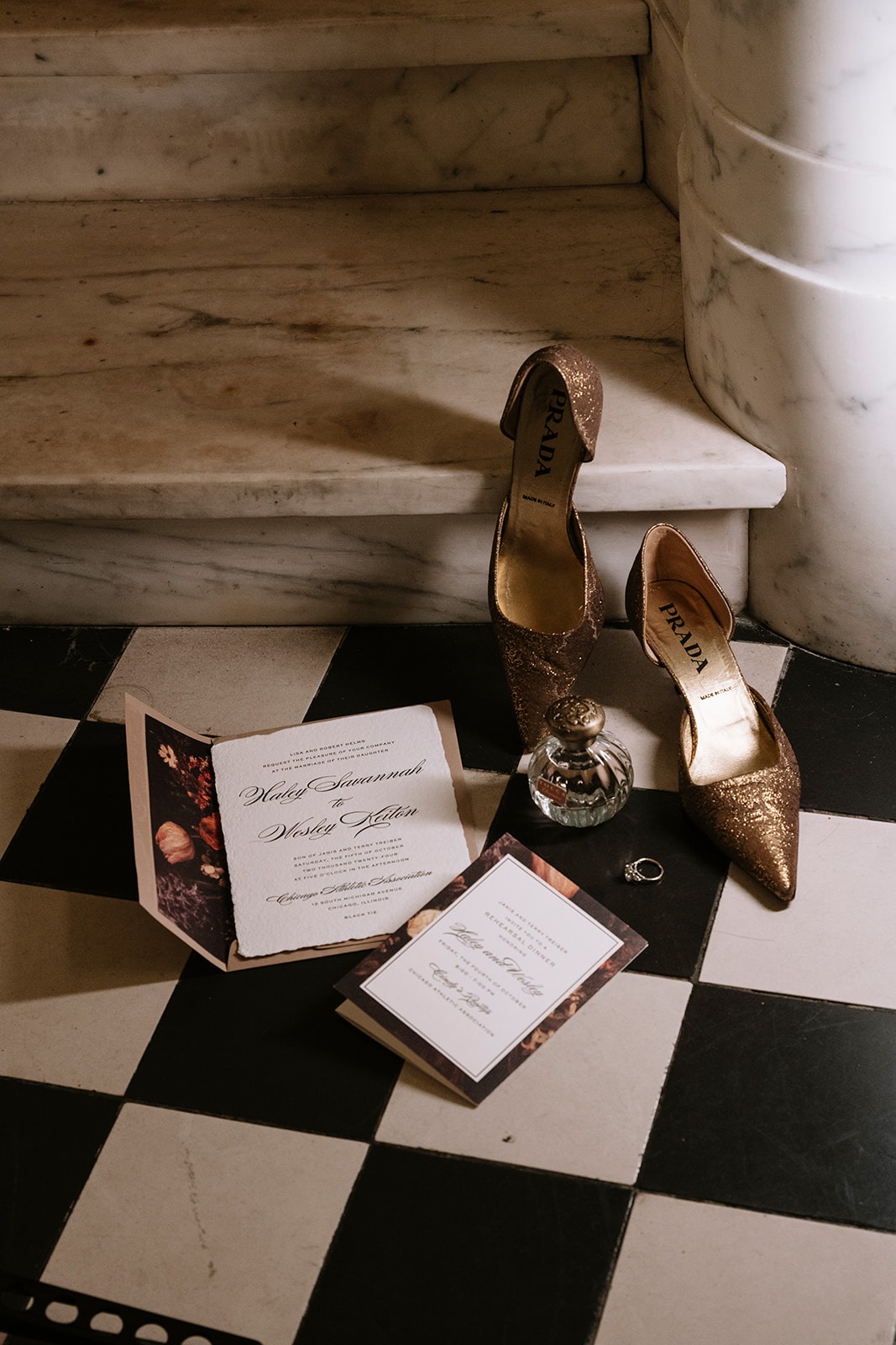 Wedding invitation suite, perfume bottle, engagement ring, and glittering gold Prada heels arranged on a black-and-white checkered floor at the foot of a marble staircase
