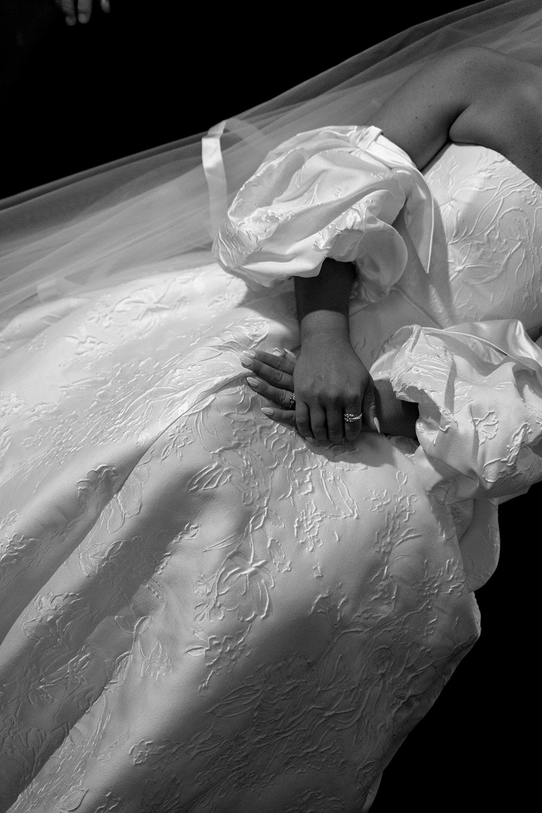 Black-and-white detail of the bride's hands resting on her floral brocade gown with puffed sleeves and a sheer veil