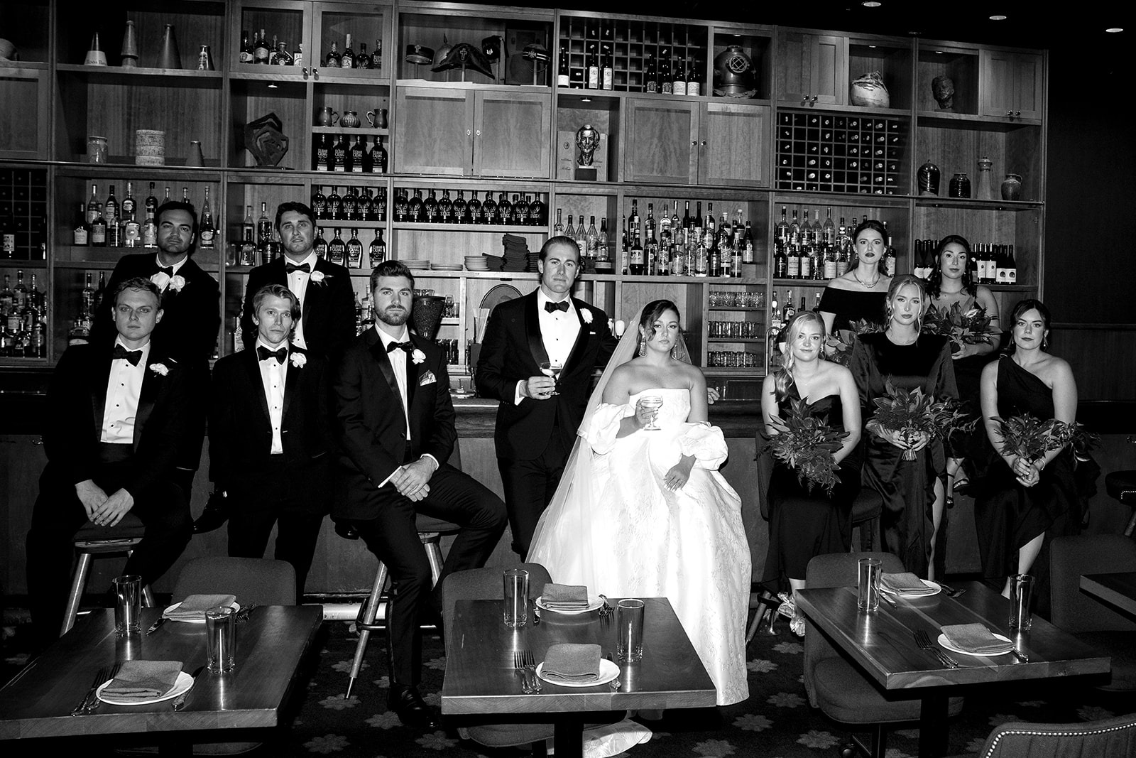 Black-and-white portrait of Haley and Wes with their bridal party in black tuxedos and black gowns, seated in front of a wood-shelved bar