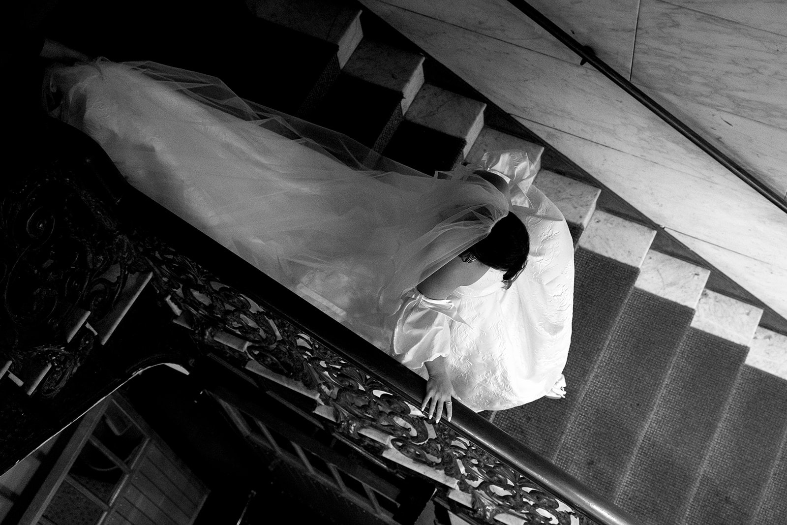 Black-and-white view from above of the bride descending a marble staircase with an ornate wrought-iron railing, her veil and train trailing behind her