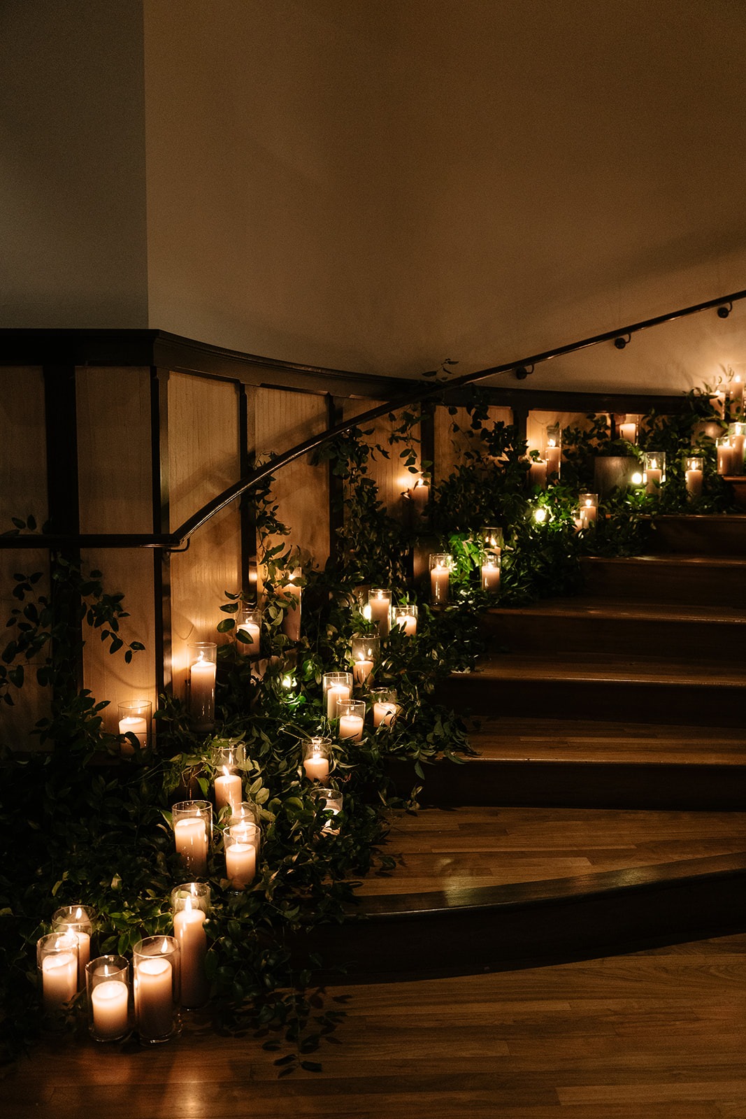 Curved wood-panelled staircase lined with trailing greenery and dozens of pillar candles in glass hurricanes