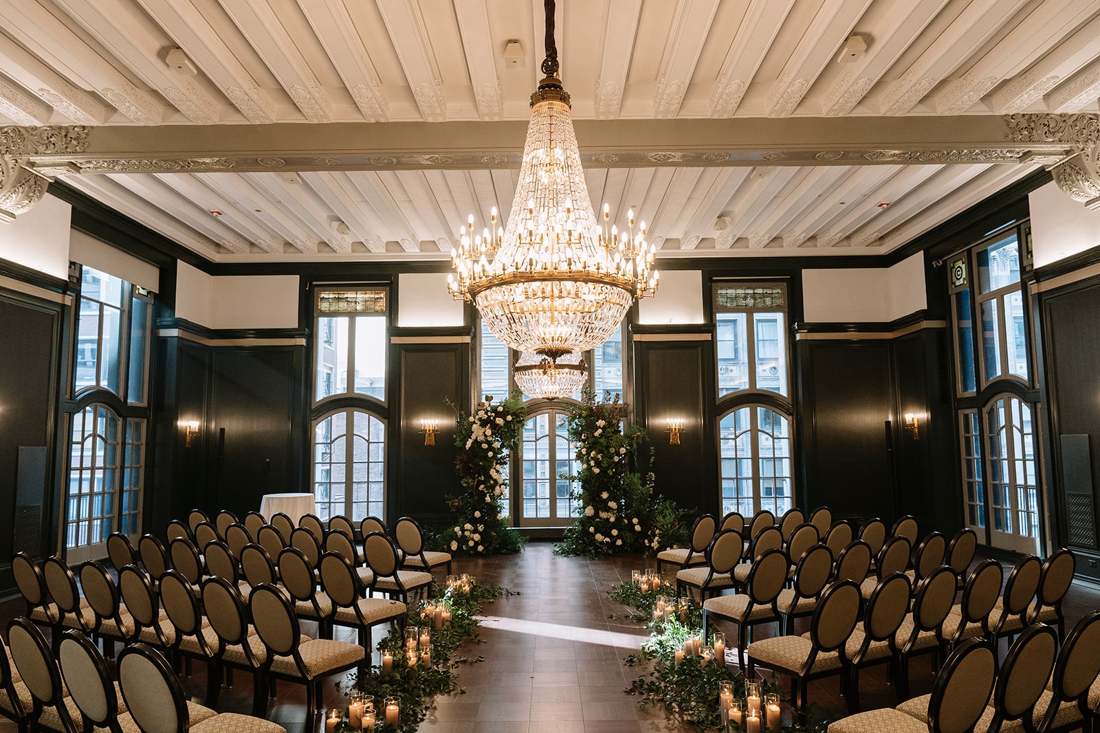 Madison Ballroom at the Chicago Athletic Association set for Haley and Wes's ceremony, with a crystal chandelier, dark wood panelling, white floral pillars framing the arched windows, and candlelit greenery lining the aisle
