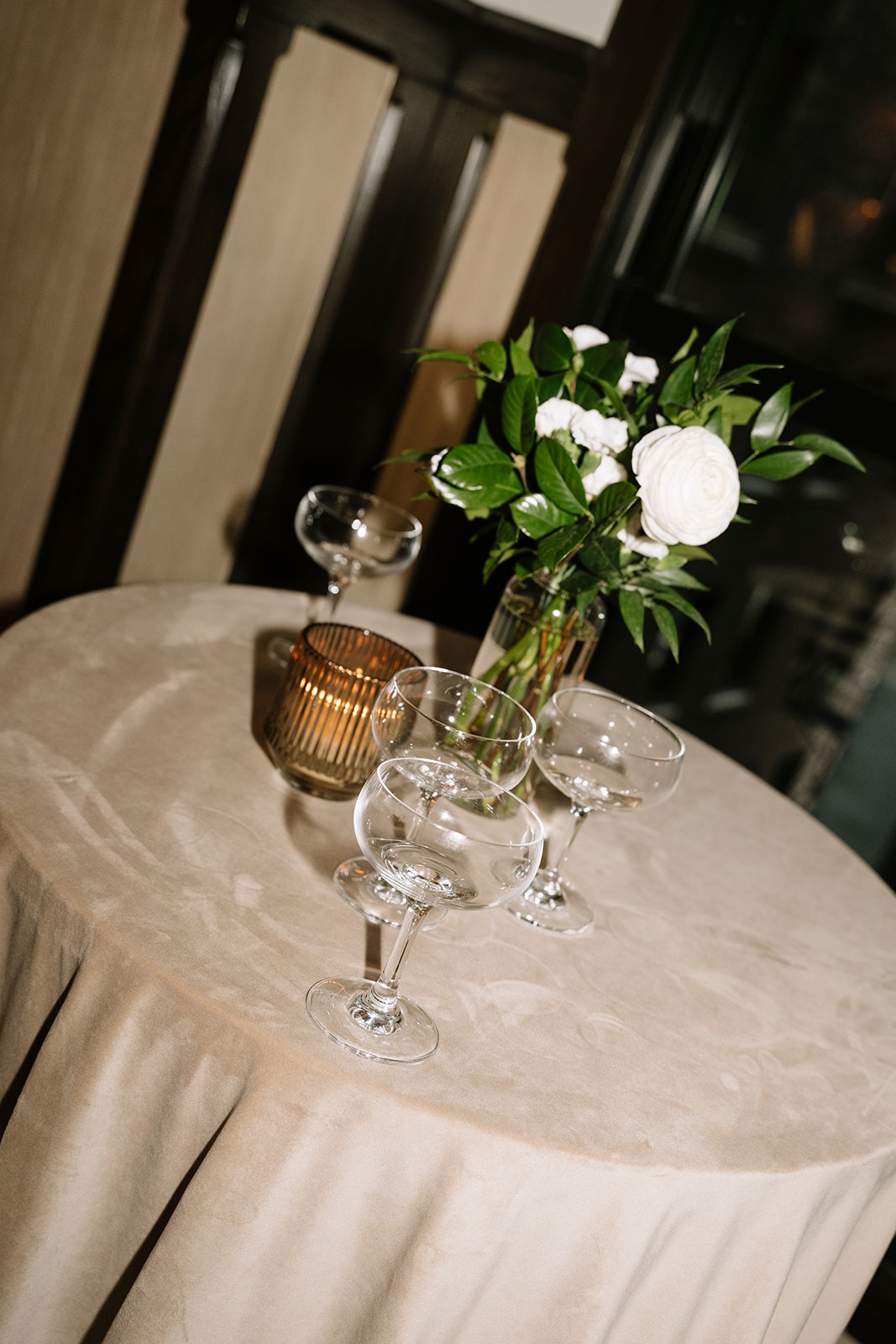 Cocktail table draped in taupe velvet with coupe glasses, an amber votive, and a small arrangement of white ranunculus and greenery