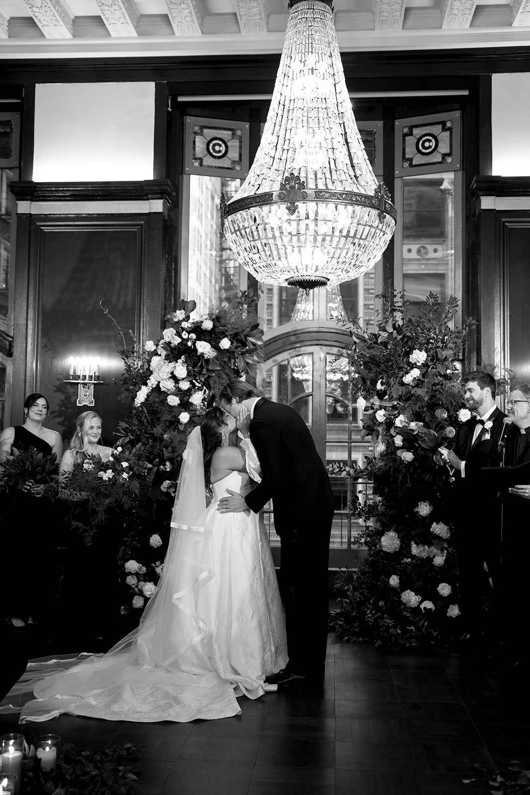 Black-and-white photograph of Haley and Wes sharing their first kiss beneath the Madison Ballroom chandelier, framed by white floral pillars as the bridal party looks on