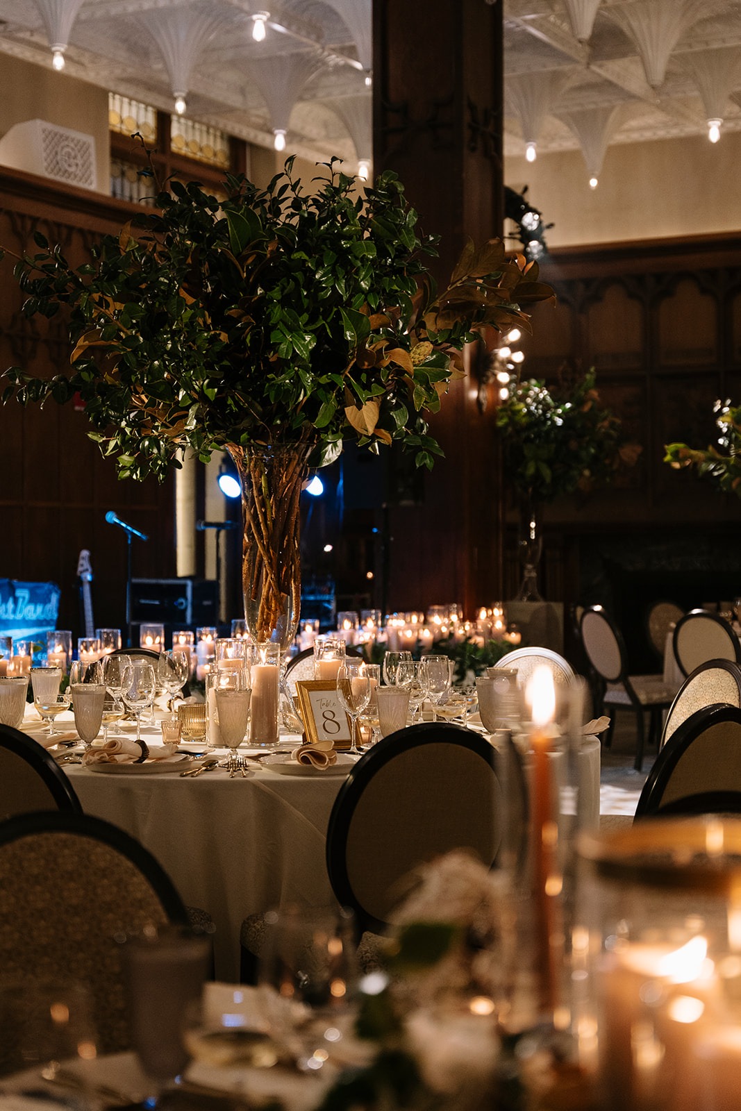 Reception table 8 with a towering greenery centerpiece, pillar candles, and a gold-framed table number beneath the White City Ballroom's ornate white ceiling, with the band stage beyond
