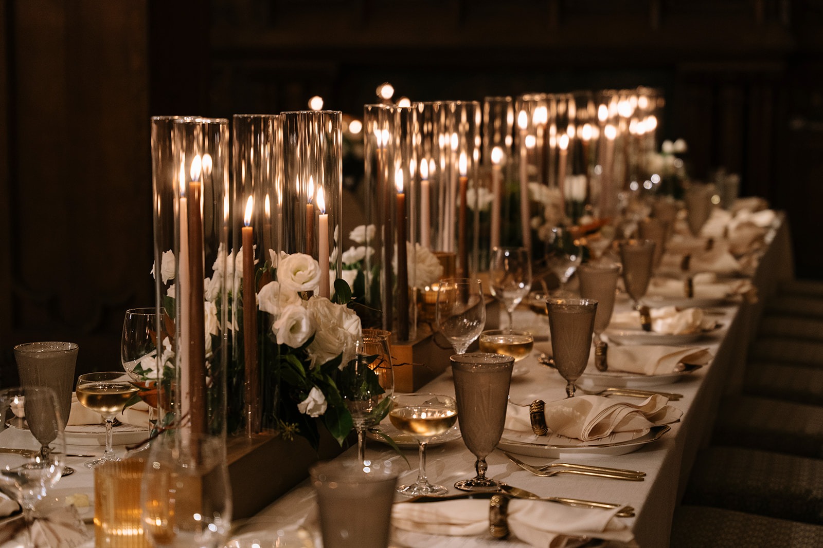 Long reception table lined with taper candles in glass cylinders, white roses, smoked-glass goblets, and gold flatware in the candlelit White City Ballroom