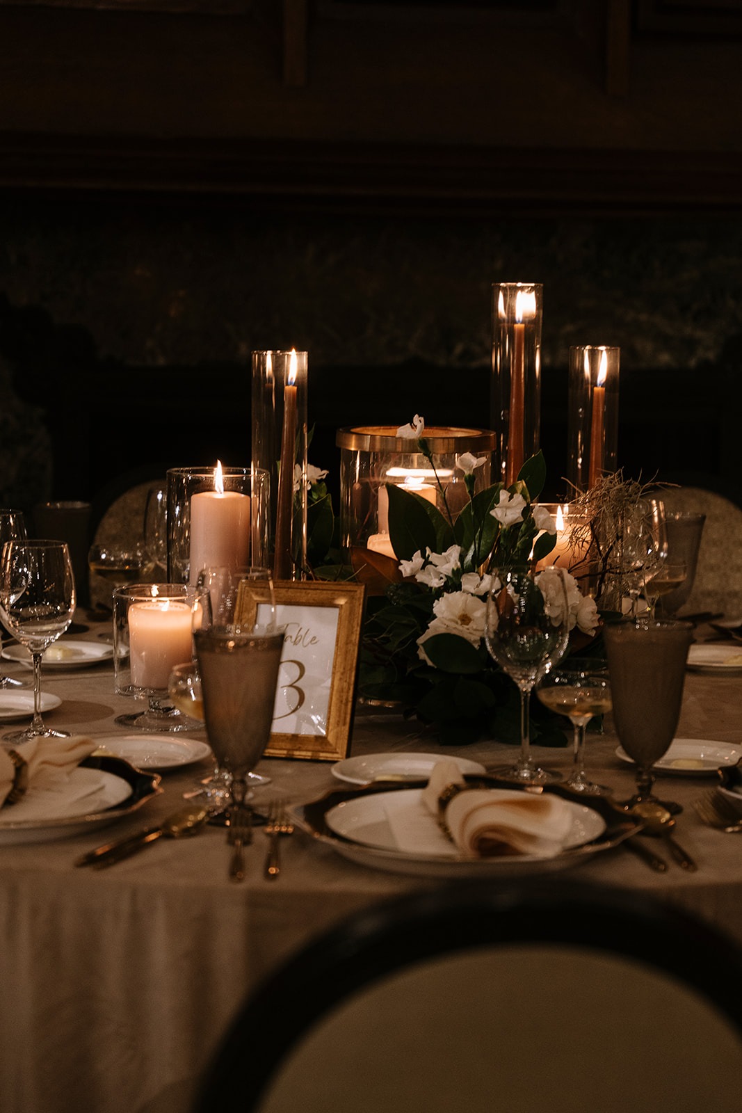 Candlelit reception table 3 with taper and pillar candles in glass hurricanes, white garden roses with magnolia leaves, a gold-framed table number, and smoked-glass goblets