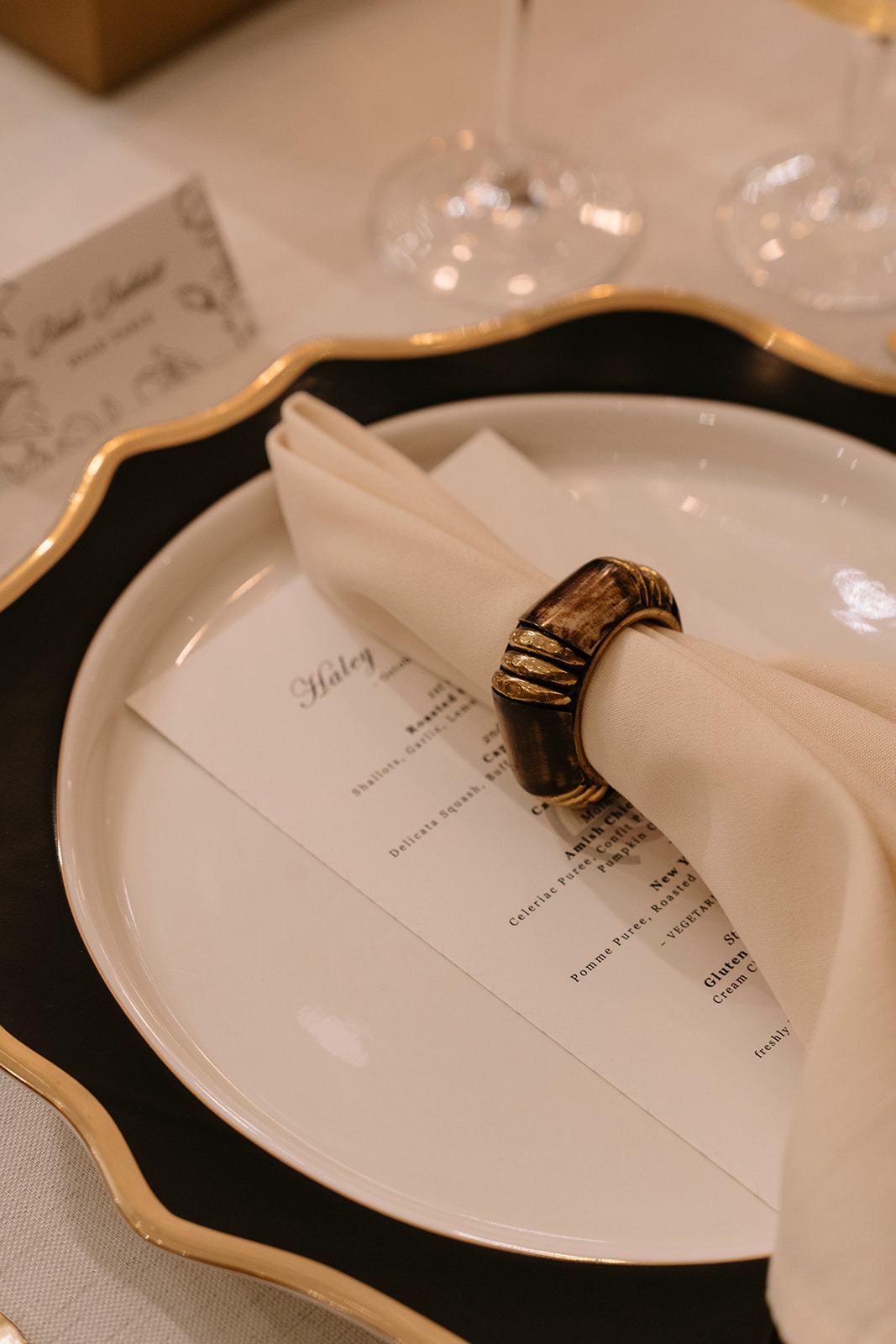 Place setting with a scalloped black-and-gold charger, white plate, printed dinner menu, and an ivory napkin held by a horn-and-brass napkin ring