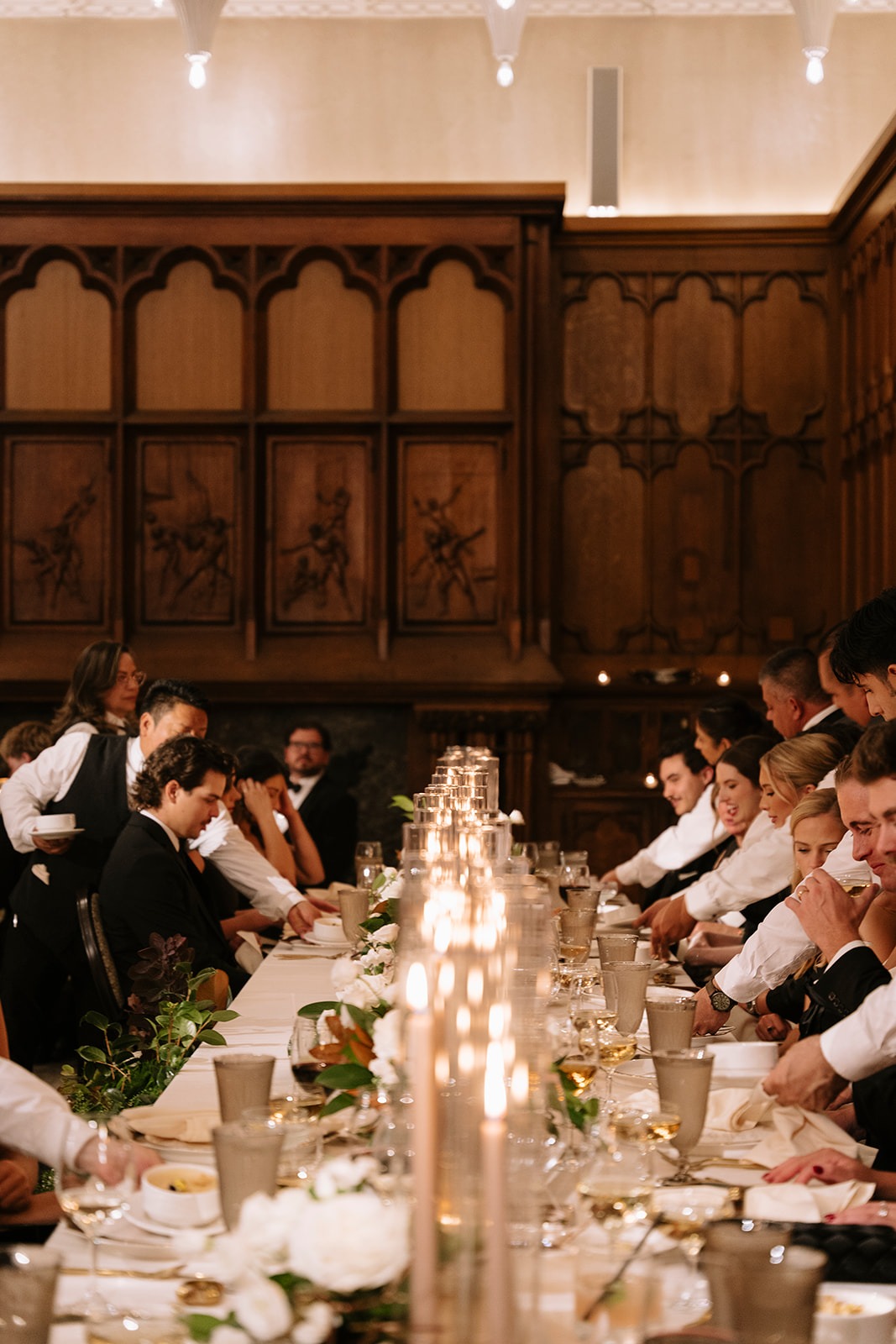 Guests dining along a long candlelit table in the wood-panelled White City Ballroom as servers present the first course