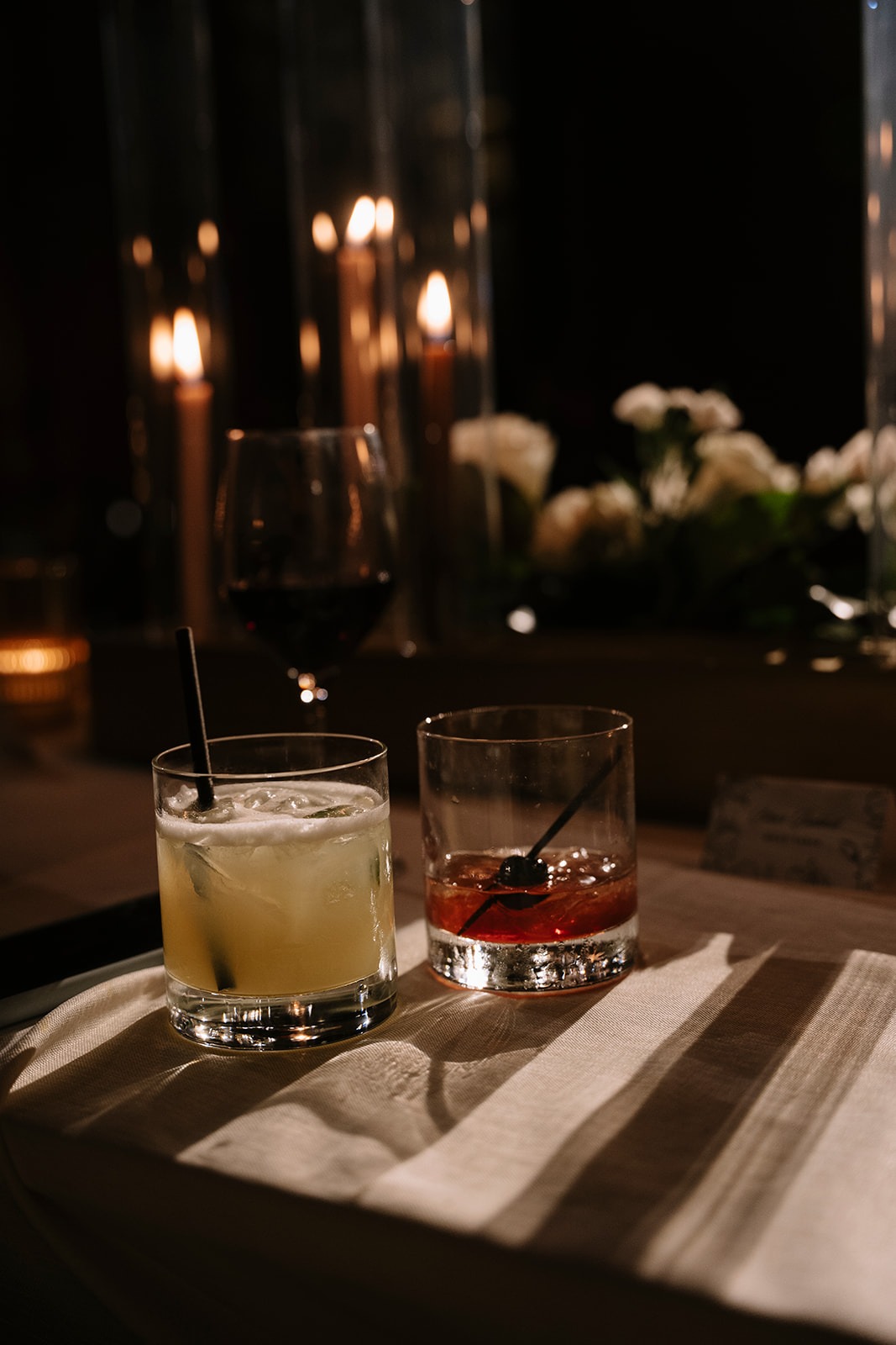 Two signature cocktails on a striped linen napkin, with a glass of red wine, taper candles, and white roses glowing behind them