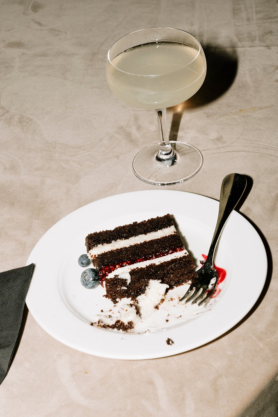 Slice of layered chocolate wedding cake with raspberry filling and fresh blueberries beside a coupe cocktail on a taupe linen