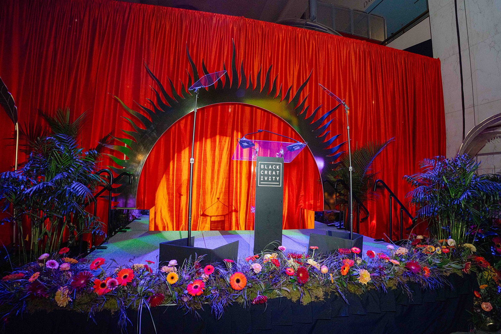 Black Creativity gala stage with a sunburst arch against red drapery, framed by palms and a bed of bright gerbera daisies, produced by Michigan Avenue Events