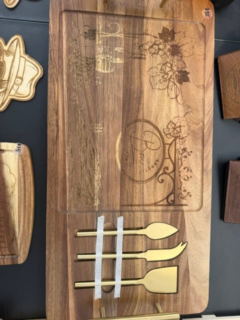 A wooden cheese board engraved with grapes, a wine bottle, and bon appetit, with gold handles and knives