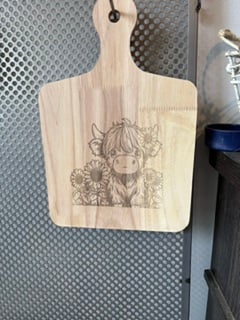 A paddle-shaped wooden cutting board engraved with a highland cow calf and sunflowers