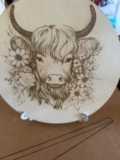 A round wooden plaque engraved with a highland cow surrounded by flowers