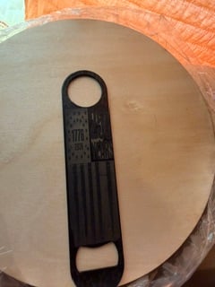 A black steel bar bottle opener engraved with an American flag and the years 1776 and 2026